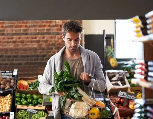 A young man walking through a grocery store aisle carrying a basket full of fresh produce, including fruits, vegetables, and dairy products, highlighting a modern grocery shopping experience.